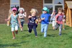 4 Jungs und 3 Mädchen rennen lachend im Sommer auf dem Rasen des Außenbereiches der Kita auf die Fotografin zu, sonniges Wetter