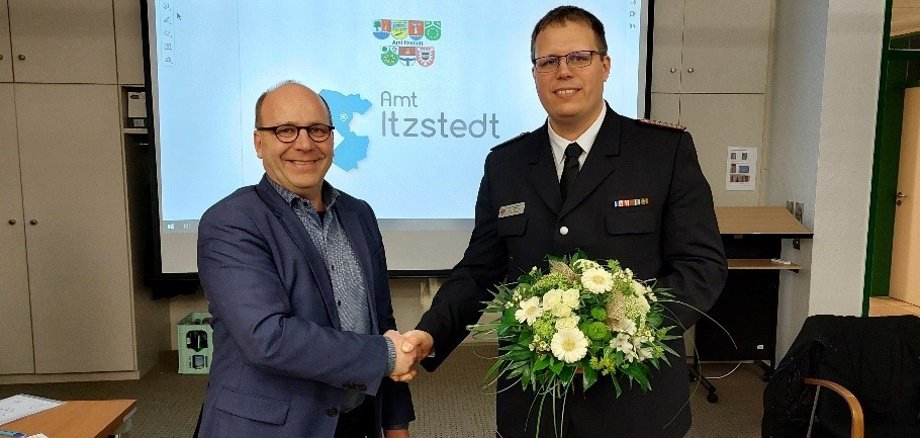 Amtsdirektor Hr. Willhoeft gratuliert Amtswehrführer Hr. Redelin, Händeschütteln, Blumenstrauß