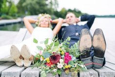Brautpaar Schuhsohlen und Brautstrauss eines mit hinter dem Kopf verschränkten Armen, sitzenden Brautpaares, welches im Hintergrund verschwommen sichtbar ist.