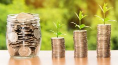 Geld Münzgeld im Einmachglas, rechts daneben 3 immer höher werdende Stapel Münzen mit wachsender Pflanze jeweils darauf